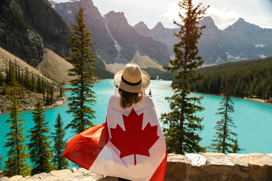 Traveller wrapped in a Canadian flag overlooking a turquoise mountain lake in Alberta, representing inter-provincial travel and insurance coverage in Canada.
